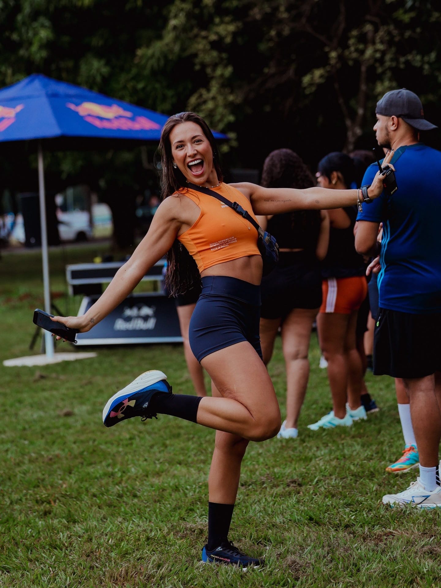 Cristina rindo em um evento esportivo ao ar livre, de top laranja, com uma perna dobrada para trás em pose de brincadeira