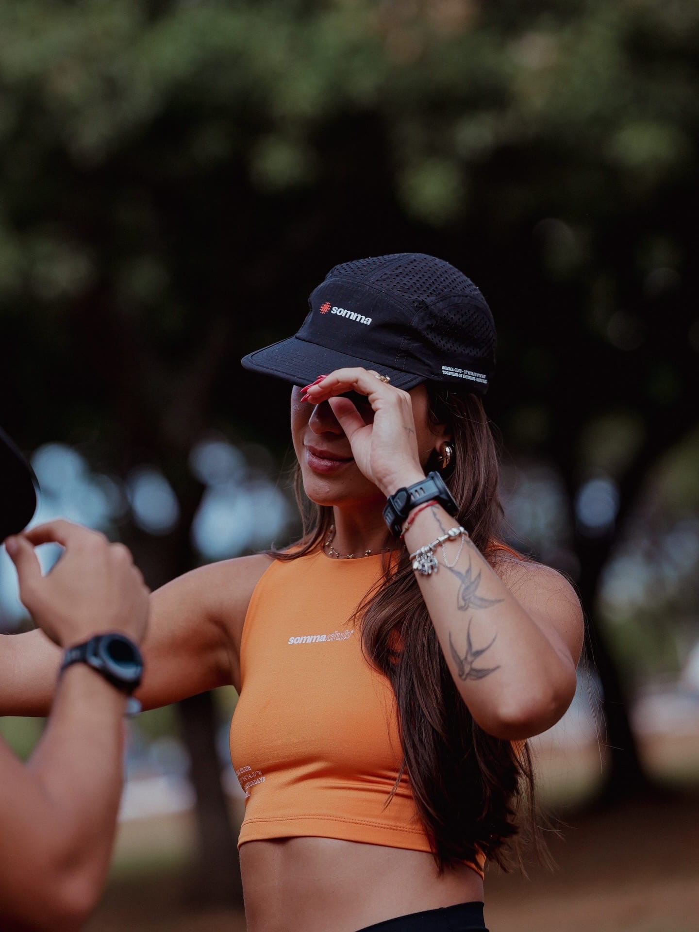Retrato esportivo de Cristina ajustando o boné preto, de top laranja, com árvores desfocadas ao fundo