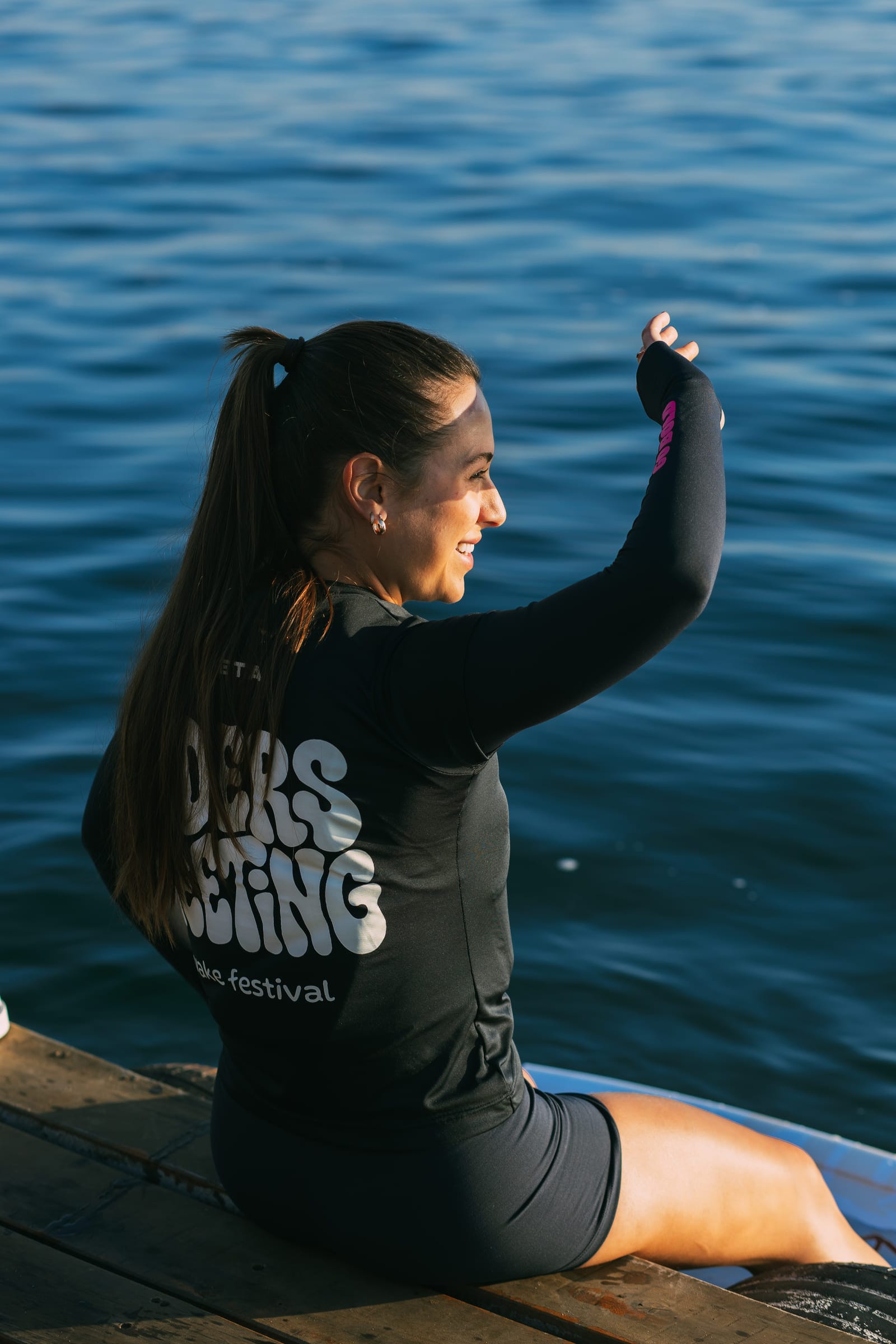 Cristina sentada no píer com a blusa do Riders Meeting wake festival, braços erguidos diante do lago